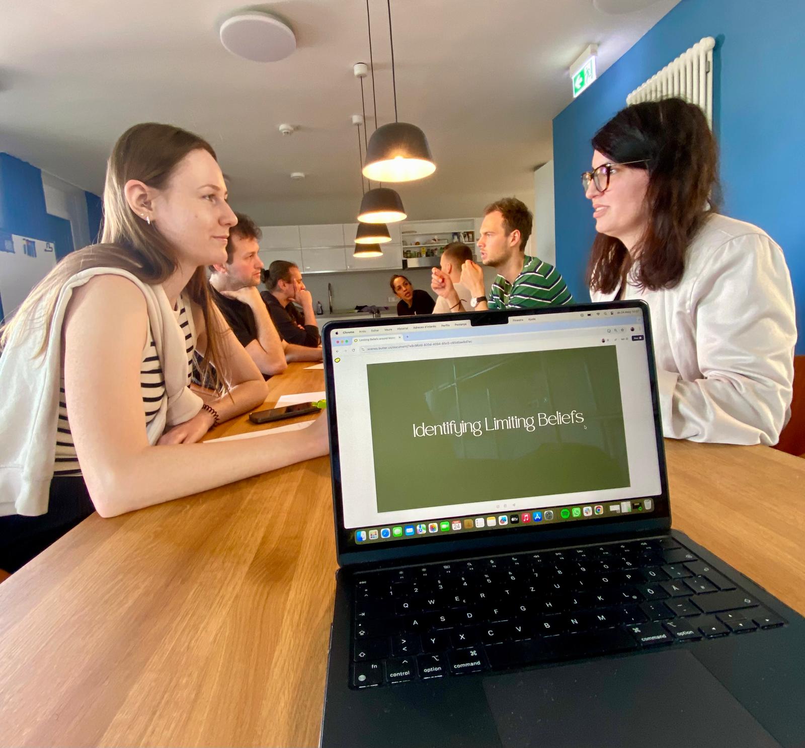 Participants seated around a table in conversation, with an open laptop in the foreground showing a slide that reads Identifying Limiting Beliefs