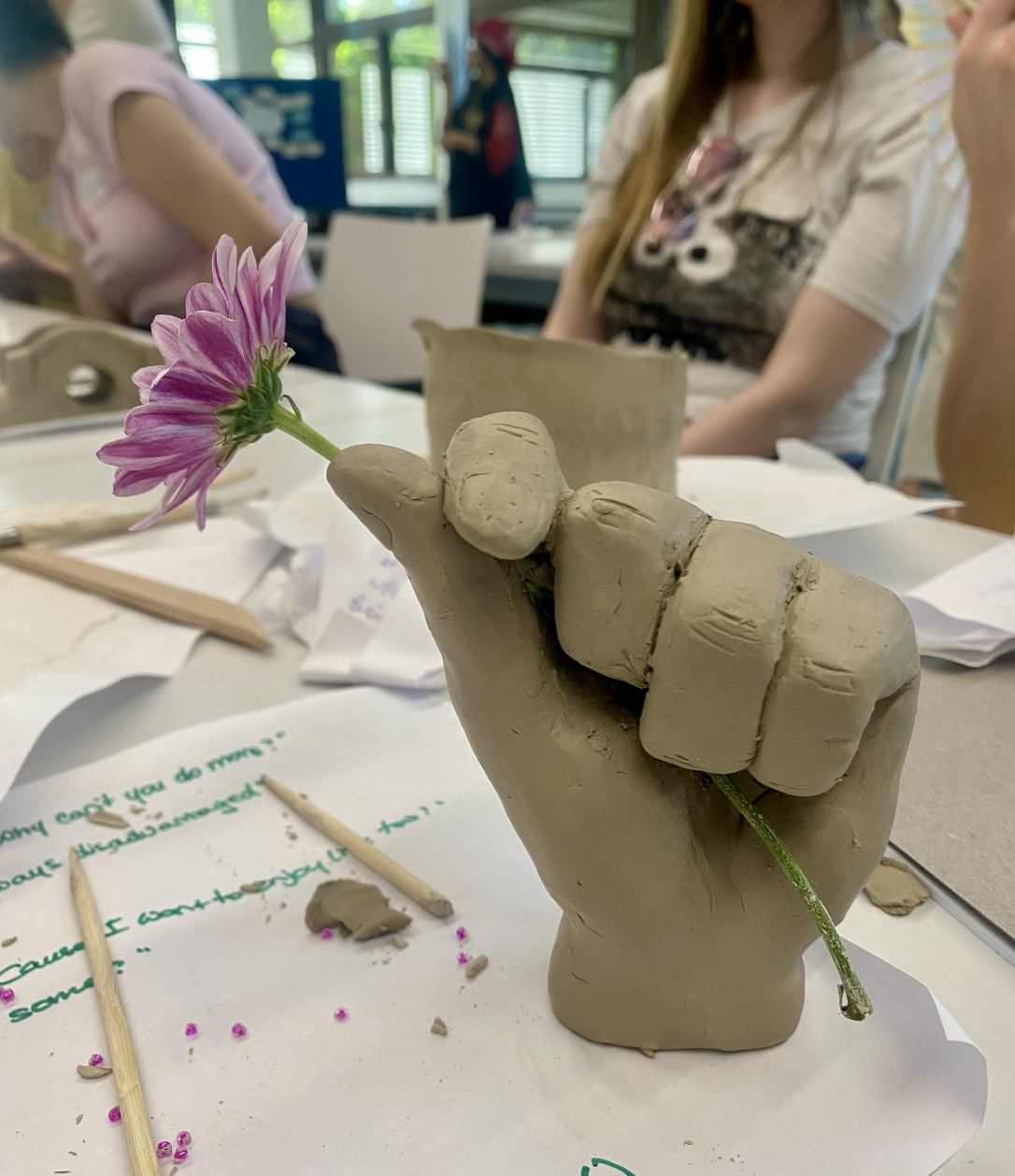 A hand sculpted from raw clay holding a pink flower, made during a workshop table session