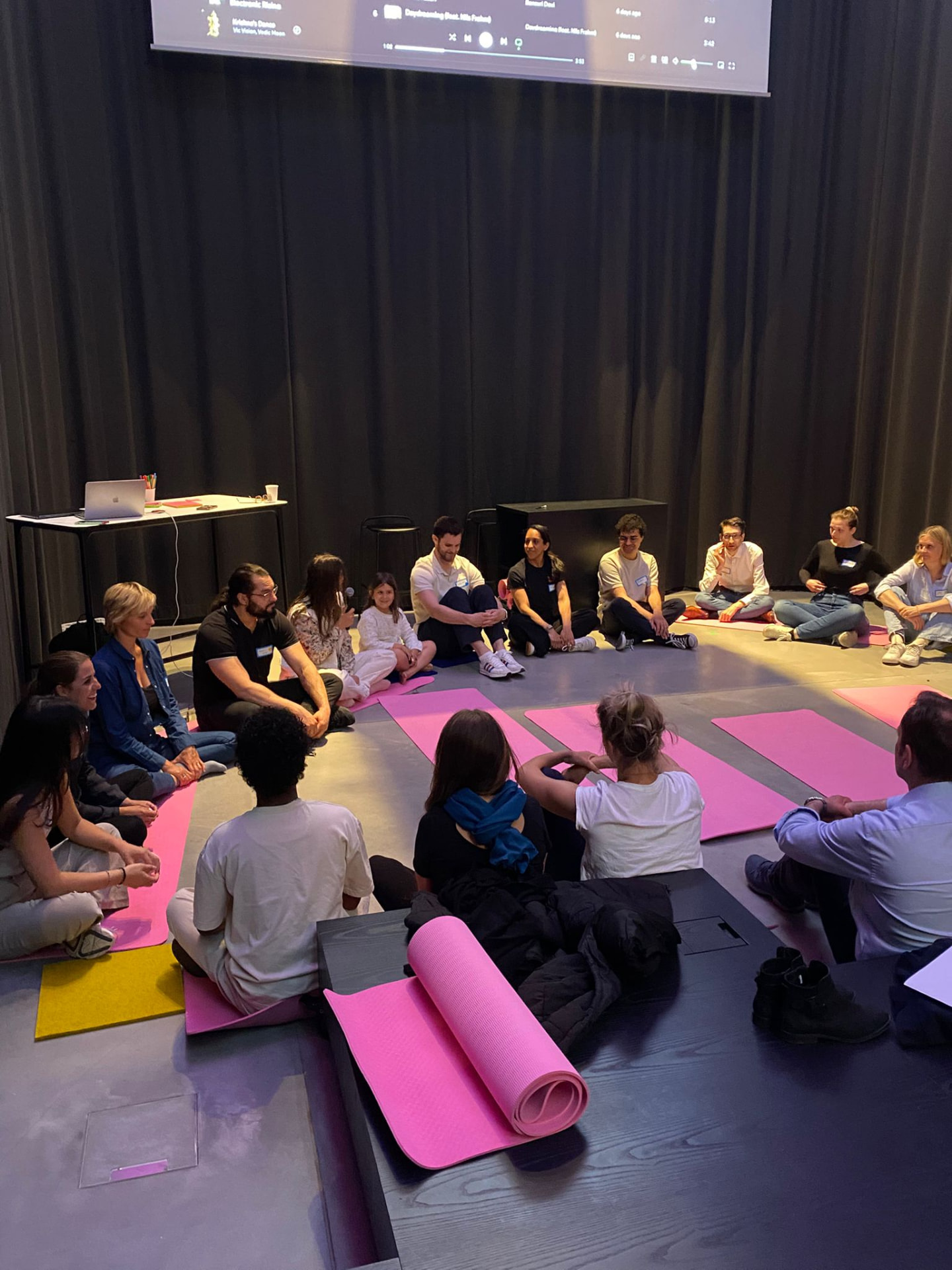 A large circle of people sitting on pink yoga mats on the floor of a darkened studio, listening as one person speaks